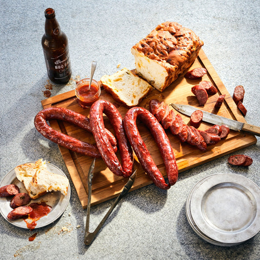 Wooden cutting board with sliced sausage on it, as well as jalapeno cheese bread, an open jar of sauce, Goode Co. BBQ sauce to the left, and a set of tin stacked plates.