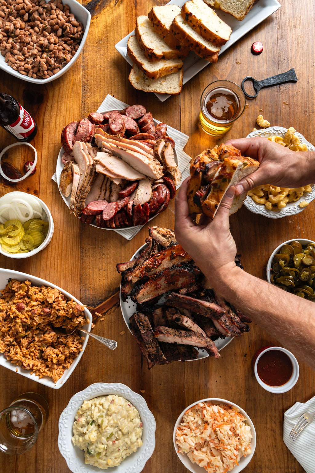 Two hands holding a brisket sandwich on jalapeno cheese bread over a Goode Co BBQ pack, including potato salad, coleslaw, sliced turkey, ribs, and pickles and onion.