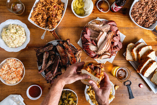 Two hands holding a brisket sandwich between two pieces of bread, over a layout of different BBQ foods.