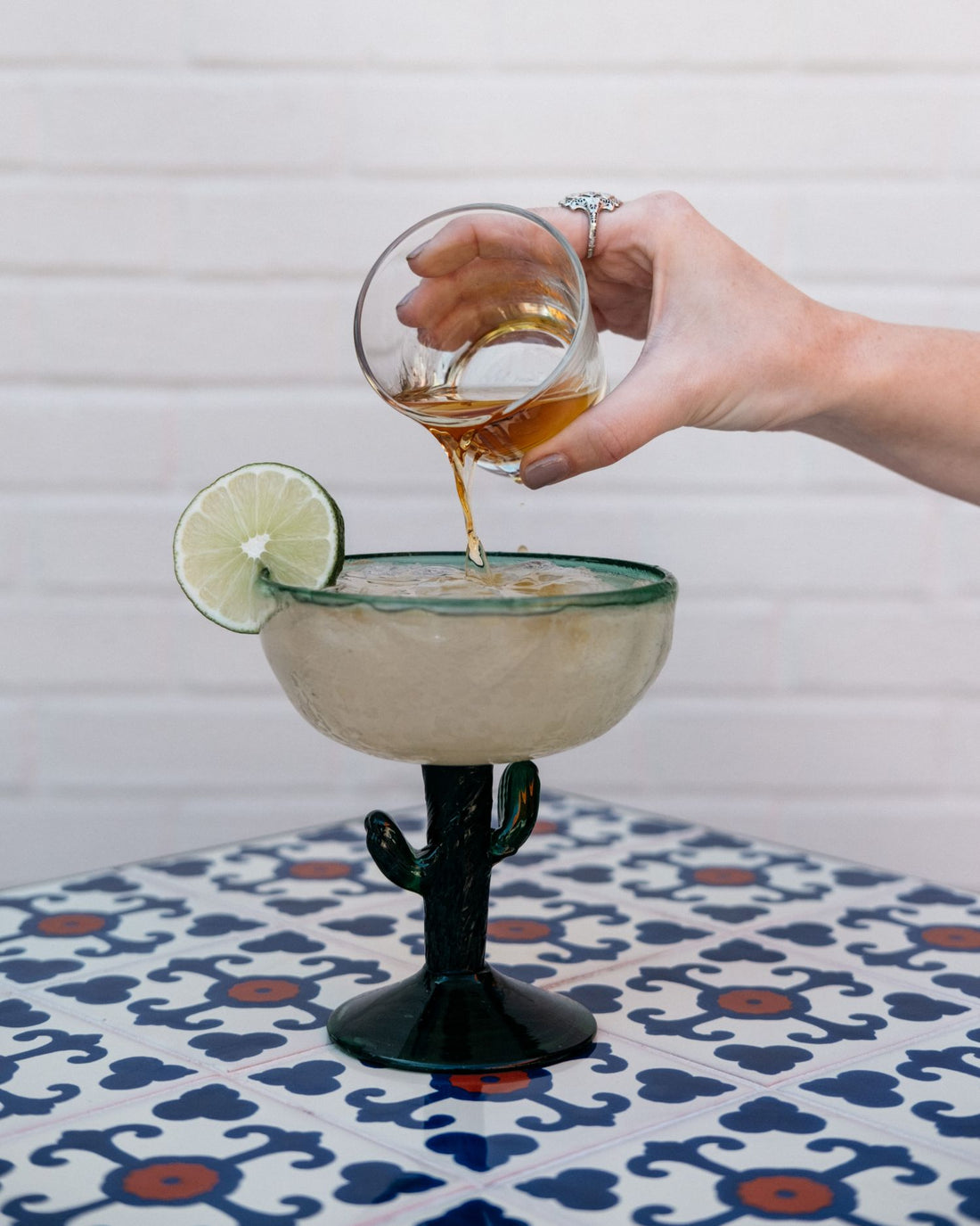 Someone with painted nails and a ring pours a shot of tequila into a full margarita glass, decorated by a lime slice.