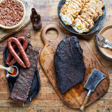 Goode Co's The Independence meal bundle, consisting of whole smoked brisket, rack of pork ribs, link of Czech sausage, link of jalapeño pork sausage, bottle Goode's Original BBQ sauce, and 2 loaves of jalapeño cheese bread.